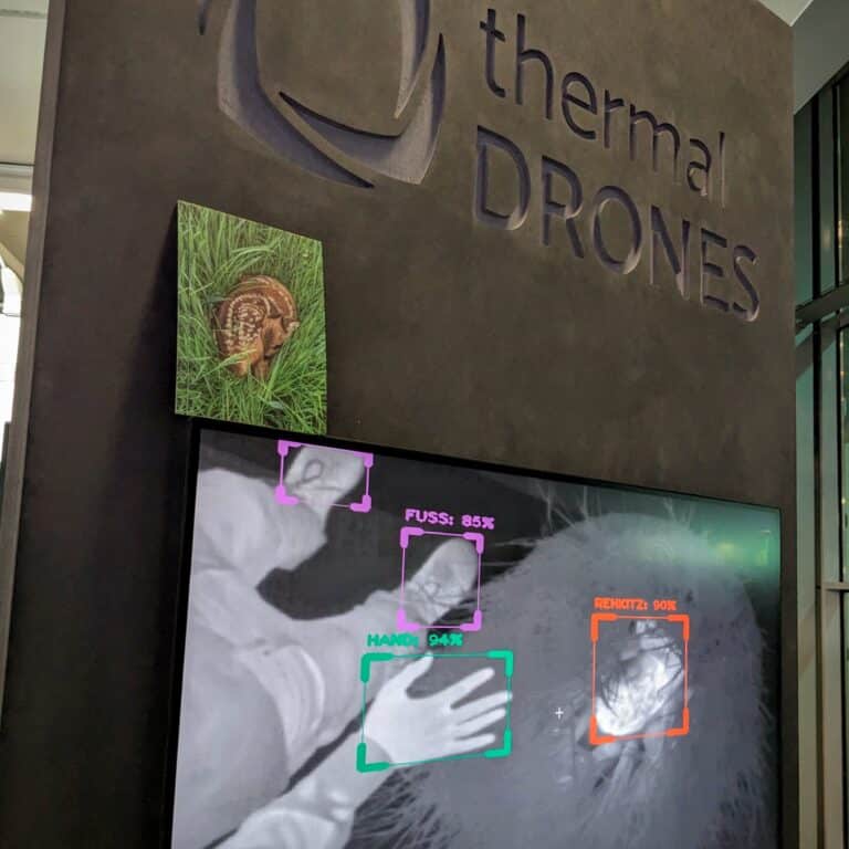 Messestand von thermal DRONES auf der Agritechnica 2023, bei welchem auf einem Bildschirm KI-basierte Objekterkennung in Echtzeit gezeigt wird. Eine Postkarte, auf der ein Rehkitz zu sehen ist, steht auf dem Bildschirm.